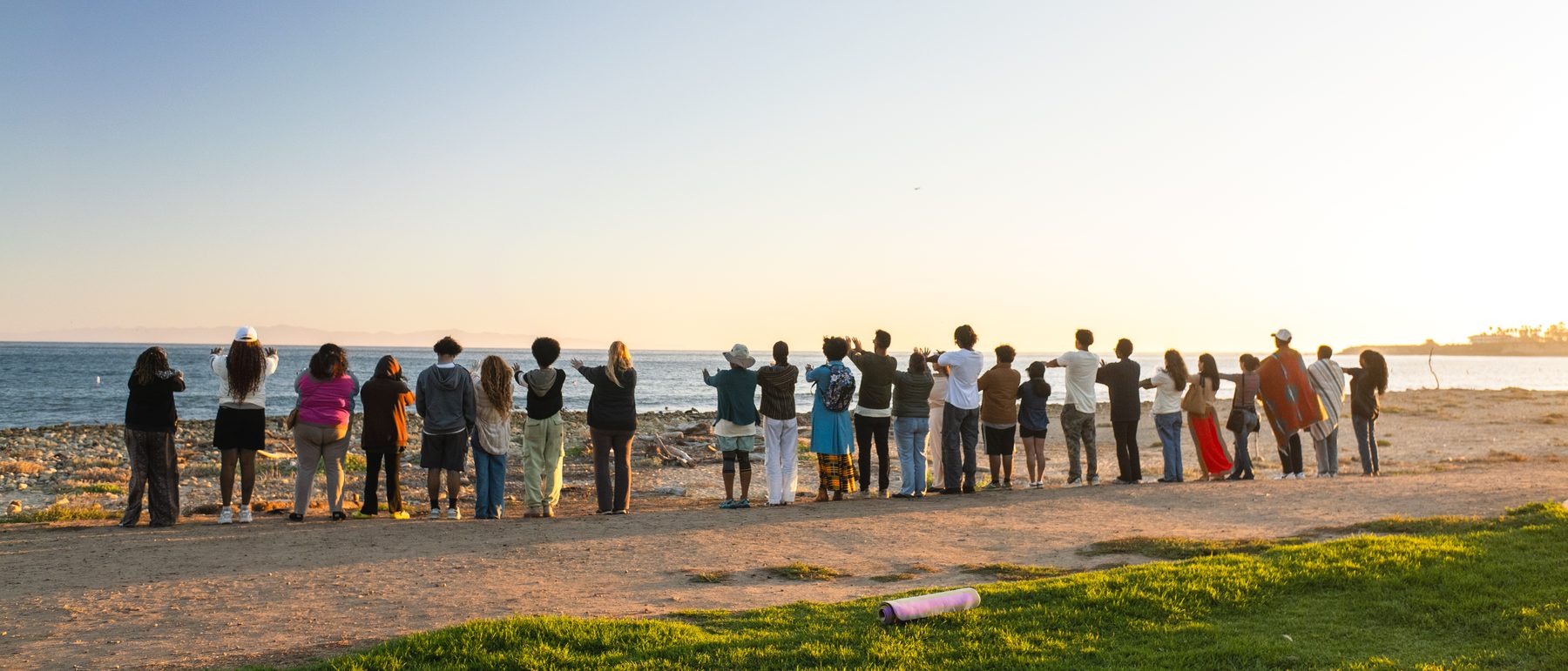 A long line of community members facing the ocean at sunset with arms outstretched, at Flourish & Flow on Goleta Beach.